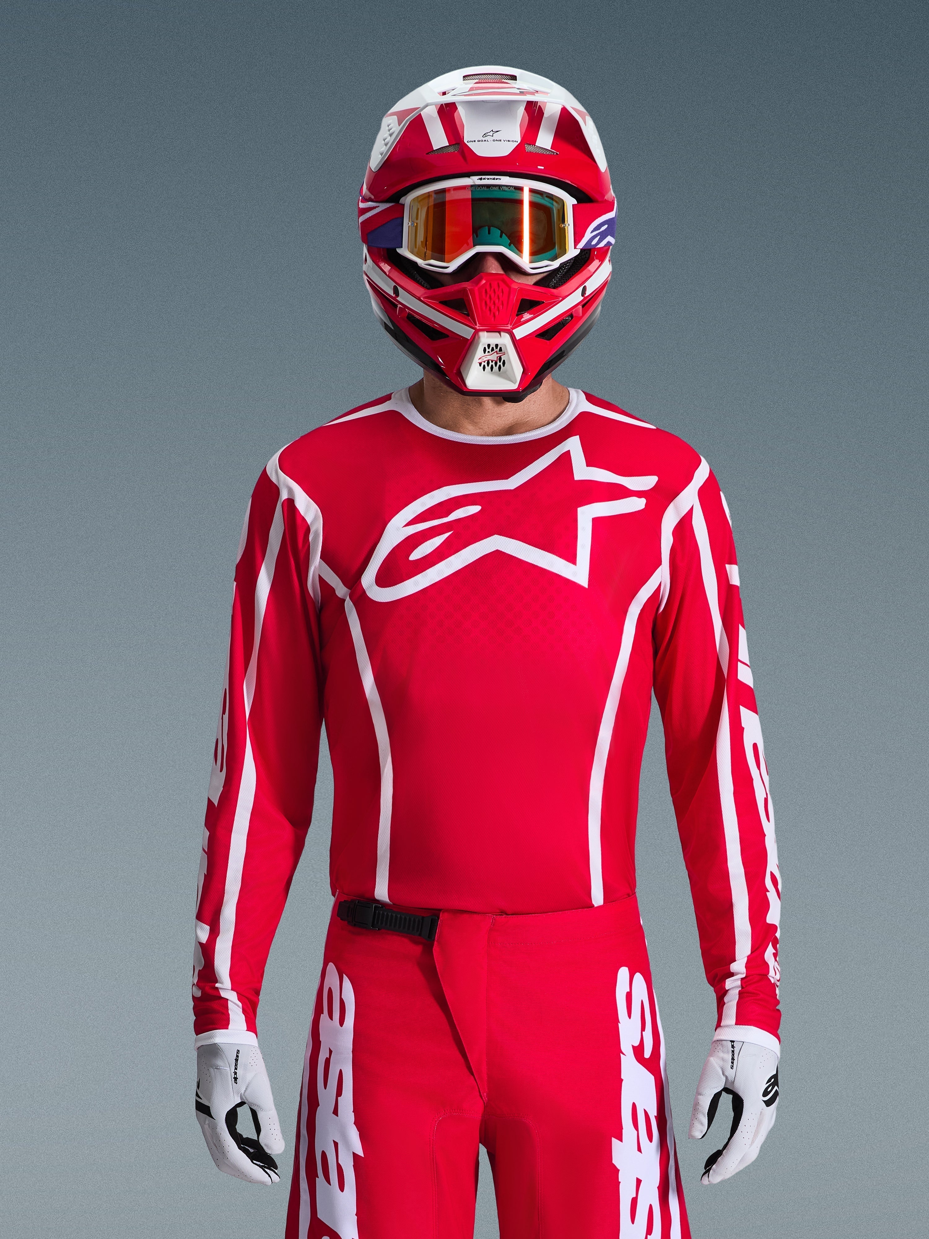 Maglia Alpinestars 2026 Fluid Apex, maglia da motocross, rossa e bianca, indossata da un pilota con casco coordinato rosso e bianco e occhiali arancioni, caratterizzata da un prominente logo bianco sul petto, vestibilità atletica, parte di un set coordinato per corse off-road