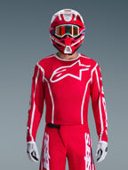 Pilota di motocross con completo Alpinestars 2026 Fluid Apex, maglia rossa con grande logo a stella bianco e profili bianchi, pantaloni rossi coordinati con accenti bianchi, casco rosso e bianco con dettagli viola e maschera con lenti arancioni, guanti bianchi e neri, outfit coordinato per corse off-road