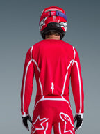 Vista posteriore di un pilota di motocross che indossa la maglia Alpinestars 2026 Fluid Apex in rosso e bianco, con motivi geometrici bianchi, abbinata a pantaloni da motocross coordinati e un casco rosso, abbigliamento completo da gara per prestazioni fuoristrada.