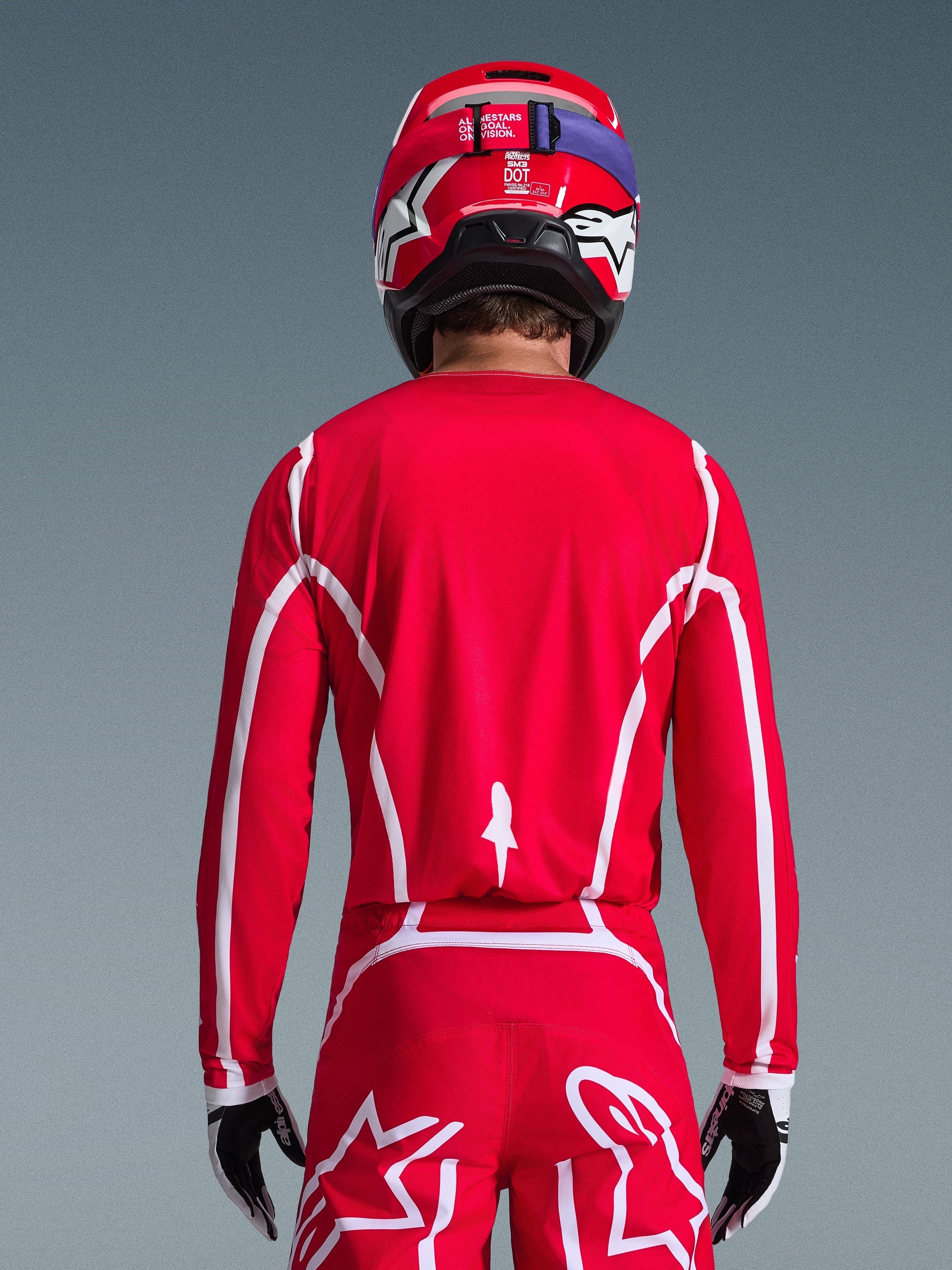 Vista posteriore di un pilota di motocross che indossa la maglia Alpinestars 2026 Fluid Apex in rosso e bianco, con motivi geometrici bianchi, abbinata a pantaloni da motocross coordinati e un casco rosso, abbigliamento completo da gara per prestazioni fuoristrada.