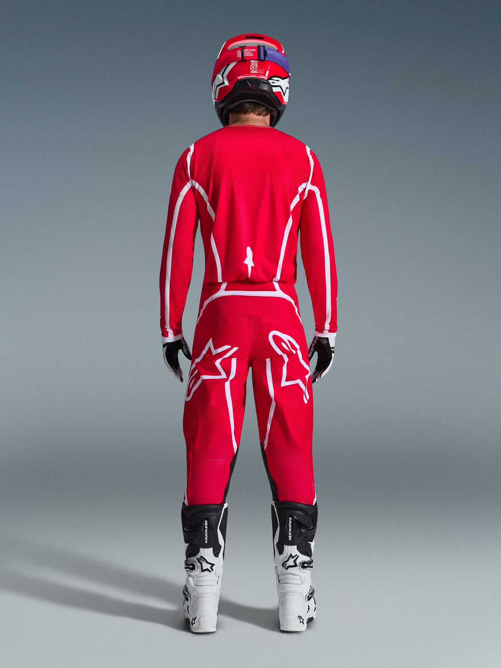 Vista posteriore di un pilota di motocross con il completo Alpinestars 2026 Fluid Apex, maglia e pantaloni rosso acceso con strisce e loghi bianchi, abbinati a stivali bianchi e neri e casco rosso, kit da corsa professionale per fuoristrada
