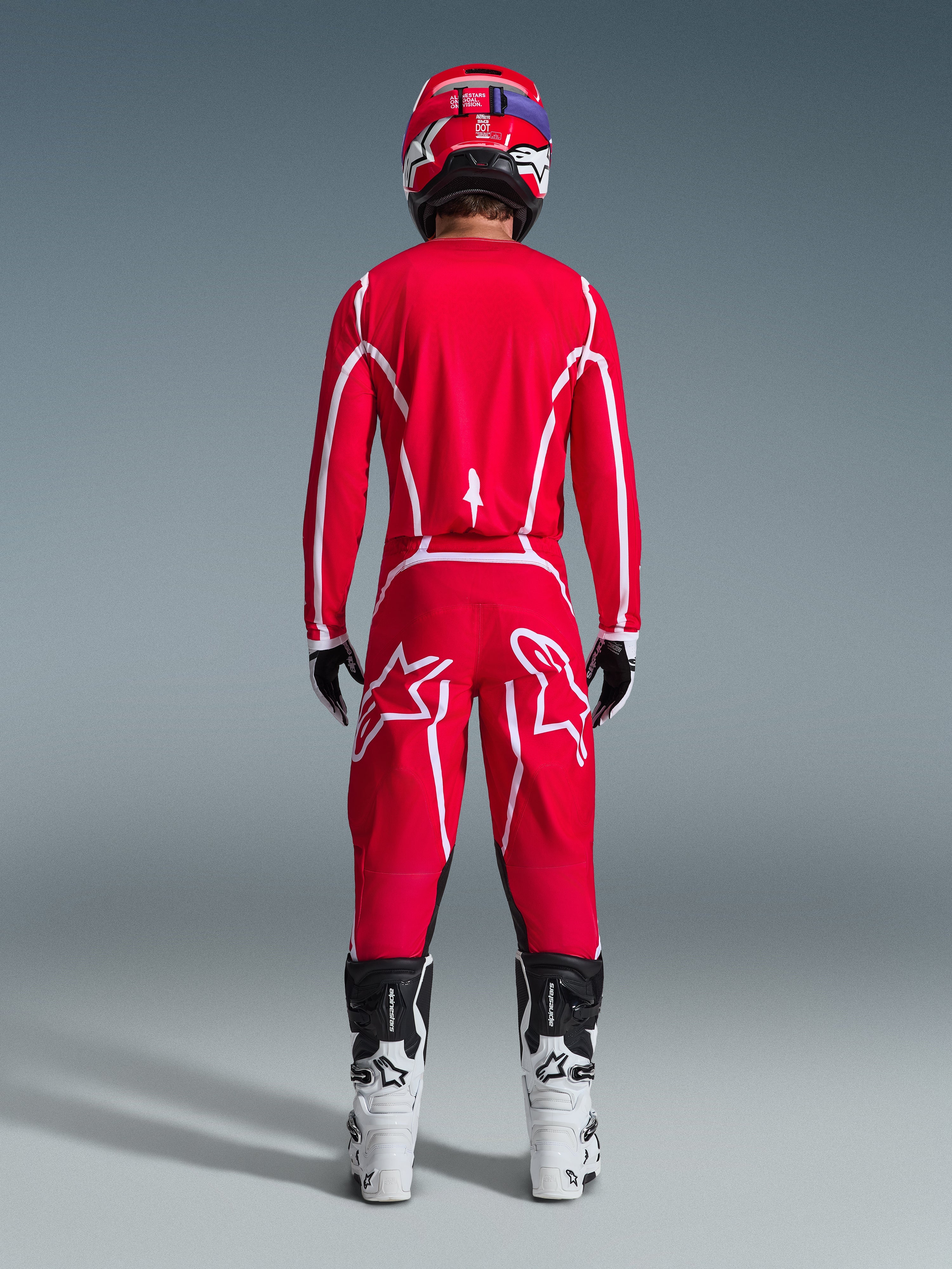 Vista posteriore di un pilota di motocross con il completo Alpinestars 2026 Fluid Apex, maglia e pantaloni rosso acceso con strisce e loghi bianchi, abbinati a stivali bianchi e neri e casco rosso, kit da corsa professionale per fuoristrada