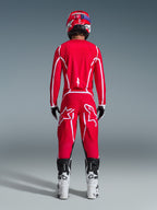 Pilota di motocross visto da dietro che indossa la maglia Alpinestars 2026 Fluid Apex in rosso e bianco, pantaloni rossi coordinati con evidenti loghi a stella bianchi, casco rosso, stivali da motocross bianchi e neri e guanti bianchi e neri, kit completo per gare off-road
