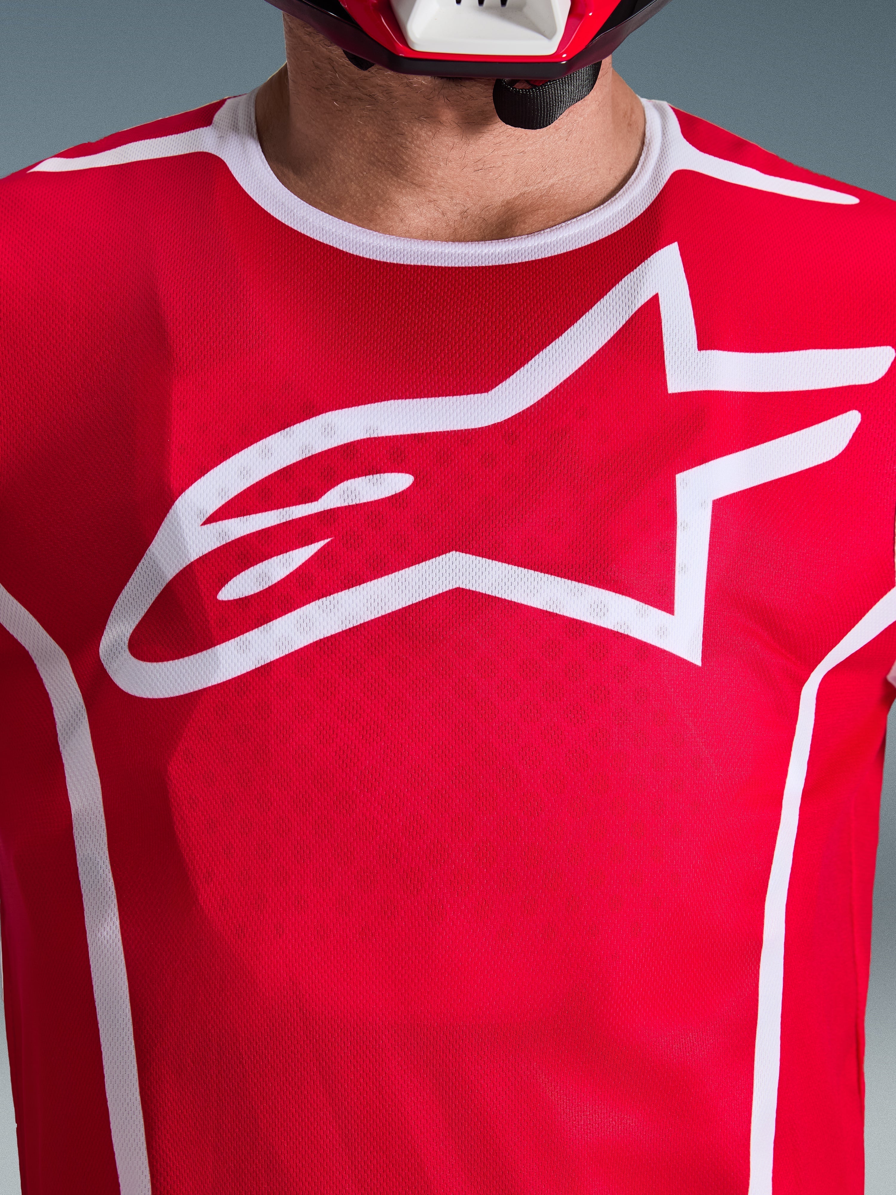 Primo piano della maglia Alpinestars Fluid Apex 2026 in rosso e bianco, caratterizzata da un grande logo Astars bianco sul petto, costruzione in mesh poly-fabric traspirante e bordo del colletto bianco, abbigliamento da motocross