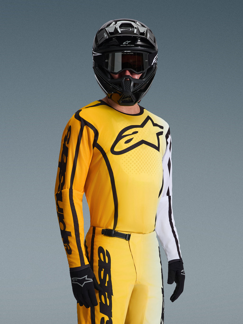 Alpinestars 2026 Fluid Apex Jersey, maglia da motocross, giallo e nero con accenti bianchi, indossata da un pilota con casco nero e occhiali, grande logo a stella nero sul petto, completo da gara off-road coordinato