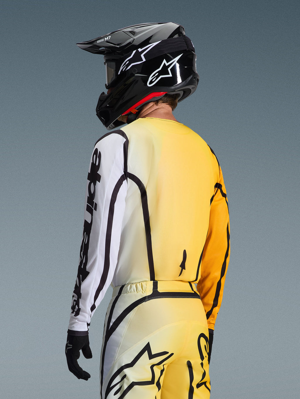 Vista posteriore di tre quarti di un pilota di motocross che indossa la maglia Fluid Apex 2026 in Giallo Nero, caratterizzata da un design sfumato dal giallo al bianco, logo Alpinestars nero ben visibile sulla manica, abbinata a pantaloni coordinati e casco da motocross nero.