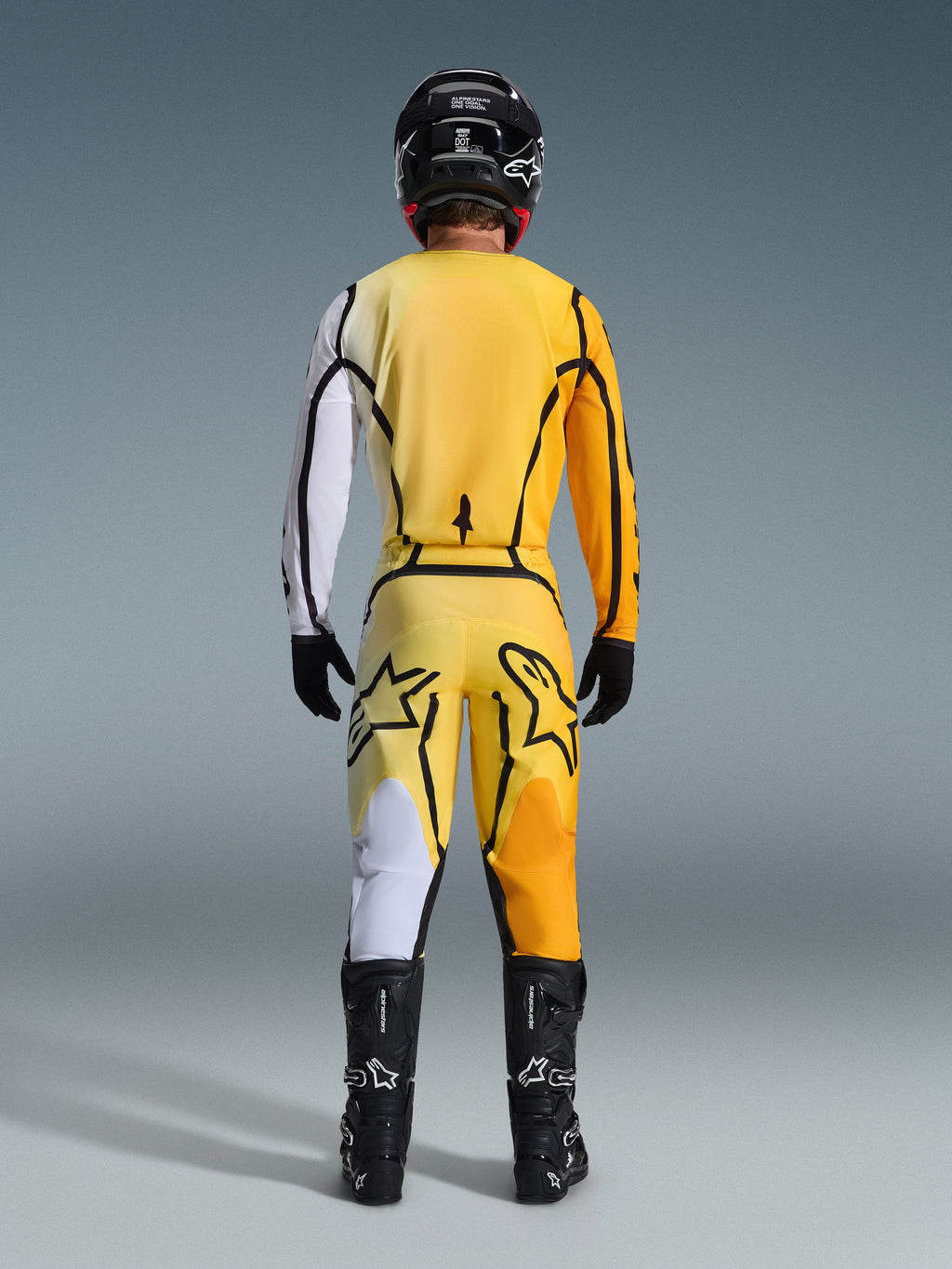 Pilota di motocross di spalle con il completo Alpinestars Fluid Apex 2026, maglia gialla con manica bianca e dettagli neri, pantaloni gialli coordinati con grandi loghi Alpinestars sul retro, casco, guanti e stivali neri, outfit coordinato per corse off-road