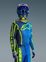 Pilota di motocross che indossa la maglia Alpinestars 2026 Fluid Apex in blu e giallo fluo, pantaloni coordinati blu e giallo neon, casco viola e giallo con occhiali blu e guanti coordinati, abbigliamento completo da gara fuoristrada.