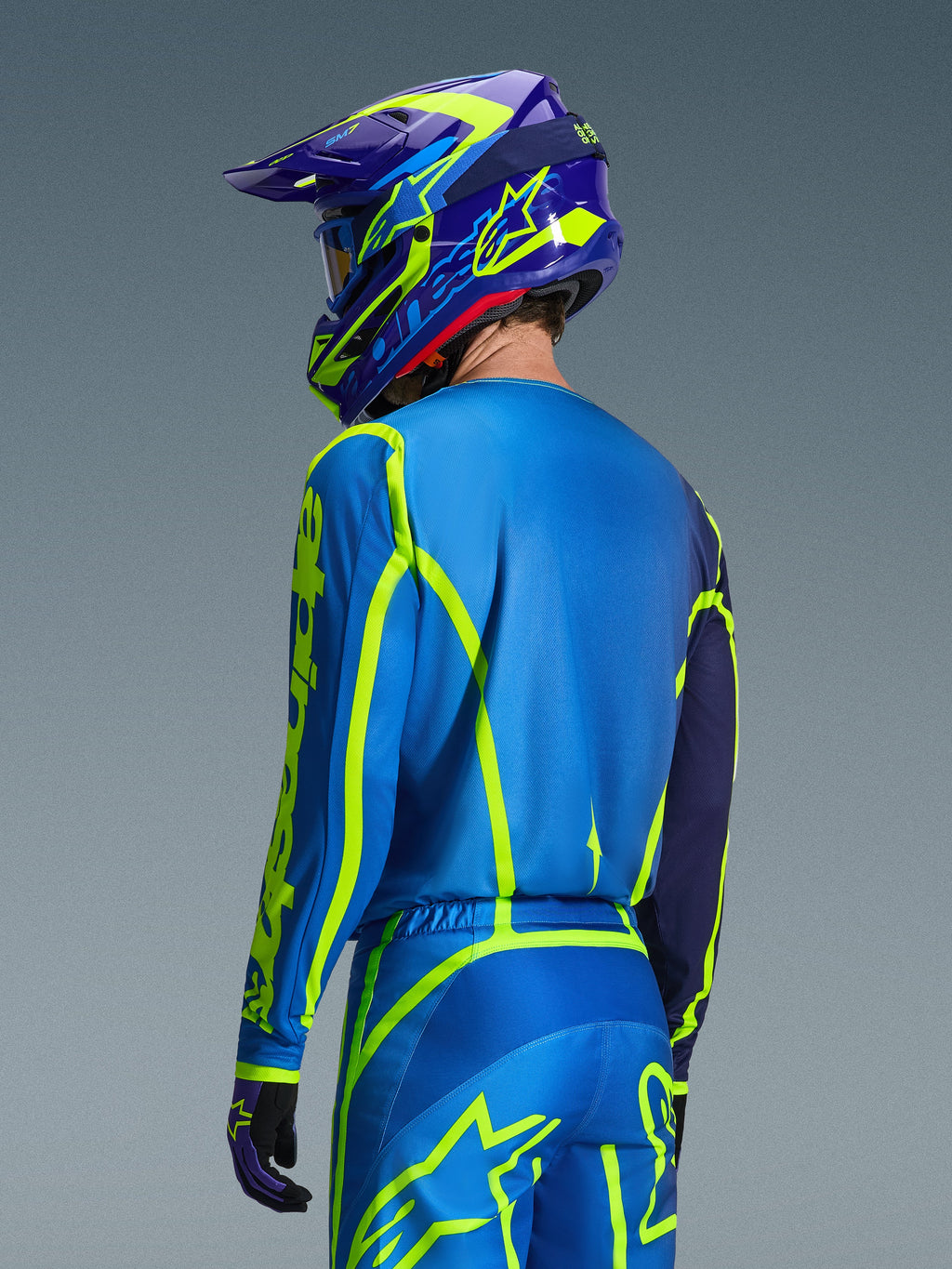 Maglia Alpinestars Fluid Apex 2026, Blu Giallo Fluo, vista posteriore del pilota che indossa pantaloni da motocross e casco coordinati, con grafiche geometriche giallo fluorescente e vestibilità atletica rilassata, abbigliamento da corsa off-road coordinato