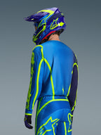 Maglia Alpinestars Fluid Apex 2026, Blu Giallo Fluo, vista posteriore del pilota che indossa pantaloni da motocross e casco coordinati, con grafiche geometriche giallo fluorescente e vestibilità atletica rilassata, abbigliamento da corsa off-road coordinato