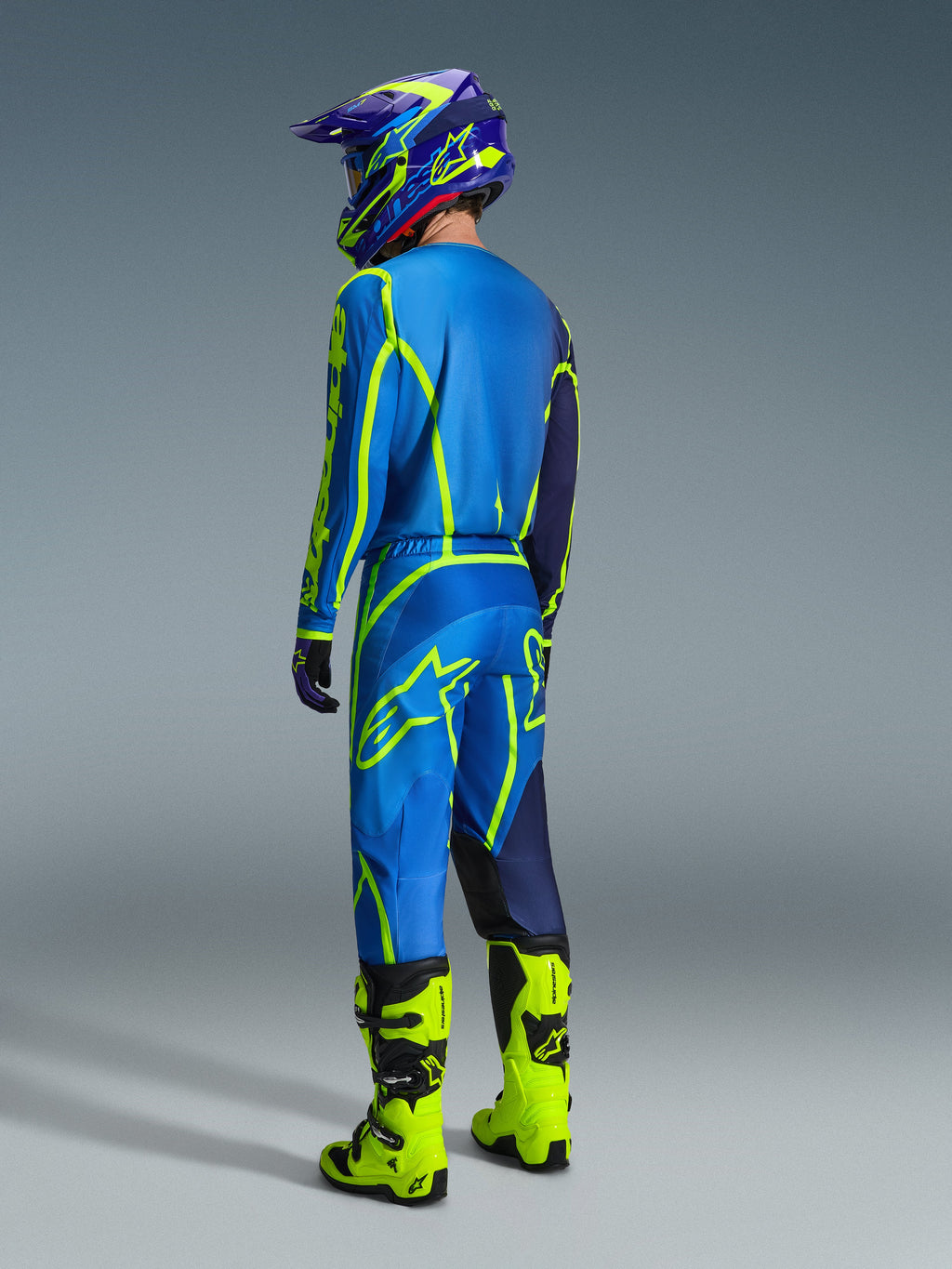 Alpinestars 2026 Fluid Apex Gear Set, pilota di motocross visto di spalle con completo blu e giallo neon, maglia e pantaloni blu con loghi giallo neon, stivali giallo neon, casco viola e blu, abbigliamento tecnico da motocross coordinato