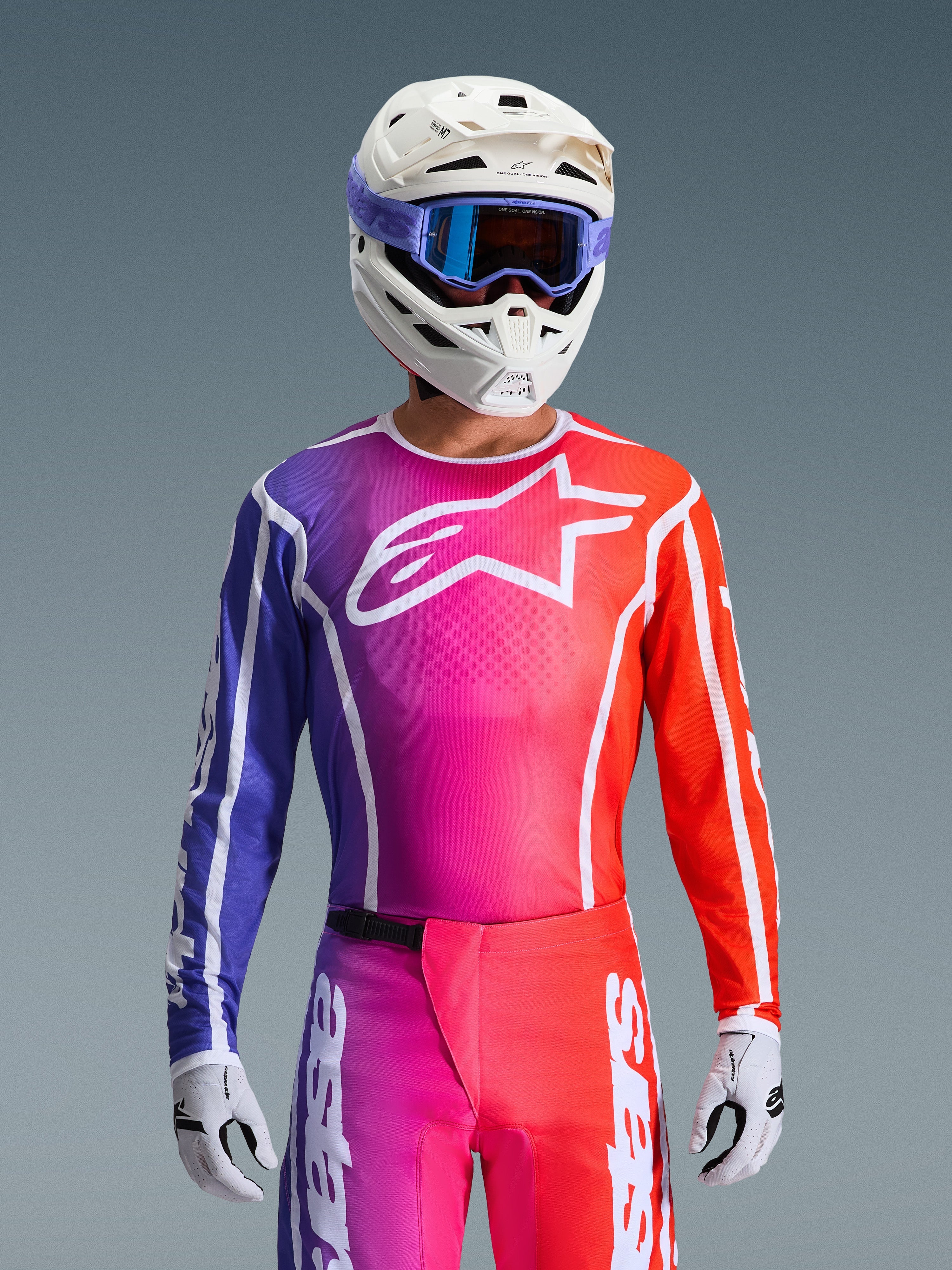 Pilota di motocross con la maglia Alpinestars Fluid Apex 2026, vivace design sfumato viola, rosa e arancione con un grande logo Alpinestars bianco sul petto, coordinata con casco bianco, maschera viola e guanti bianchi per un look da corsa fuoristrada completo.