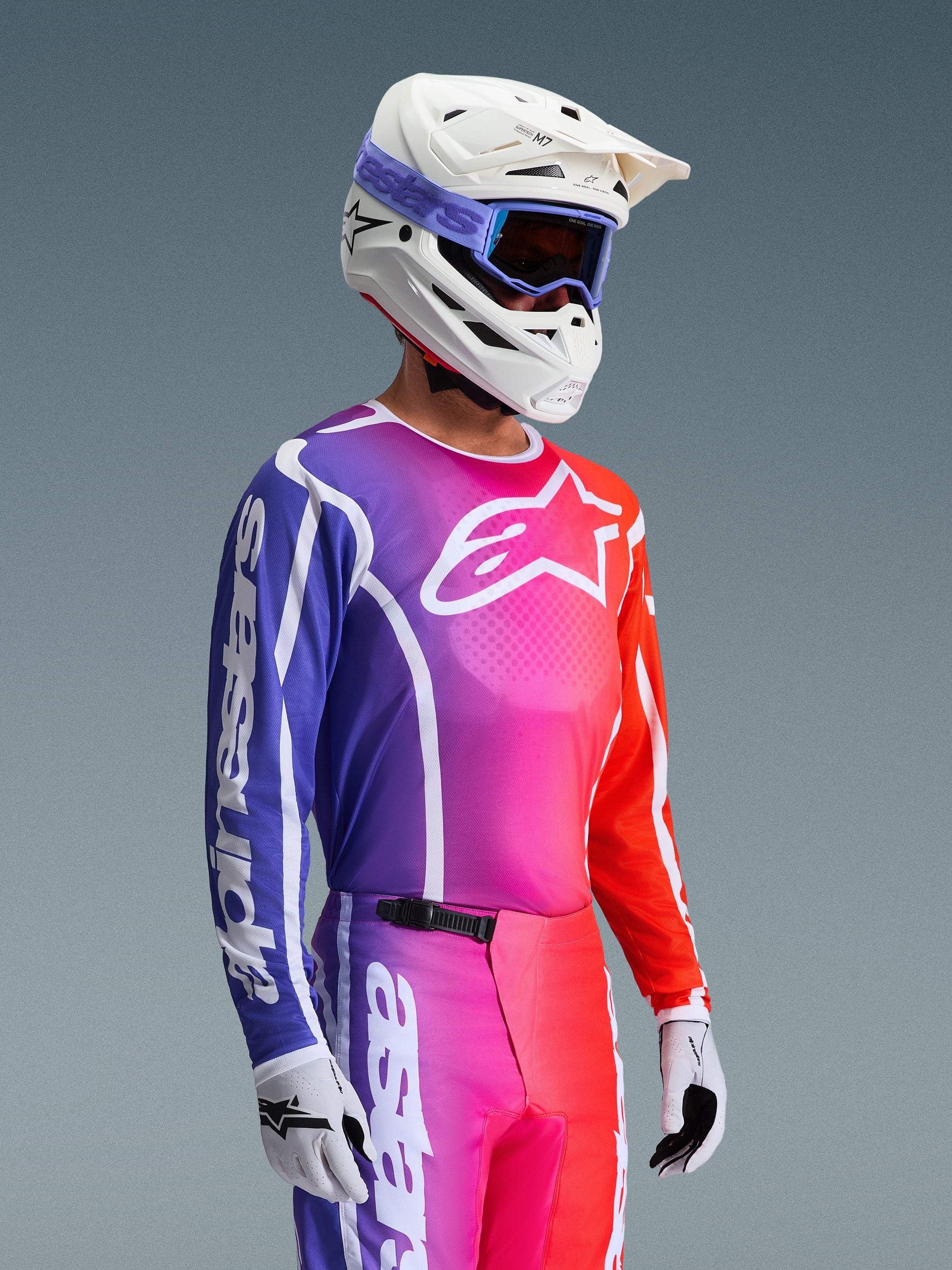 Alpinestars 2026 Fluid Apex Jersey, maglia da motocross, sfumatura vibrante viola, rosa e arancione, indossata con pantaloni coordinati, casco bianco con occhiali blu e guanti bianchi, design atletico per prestazioni e mobilità fuoristrada