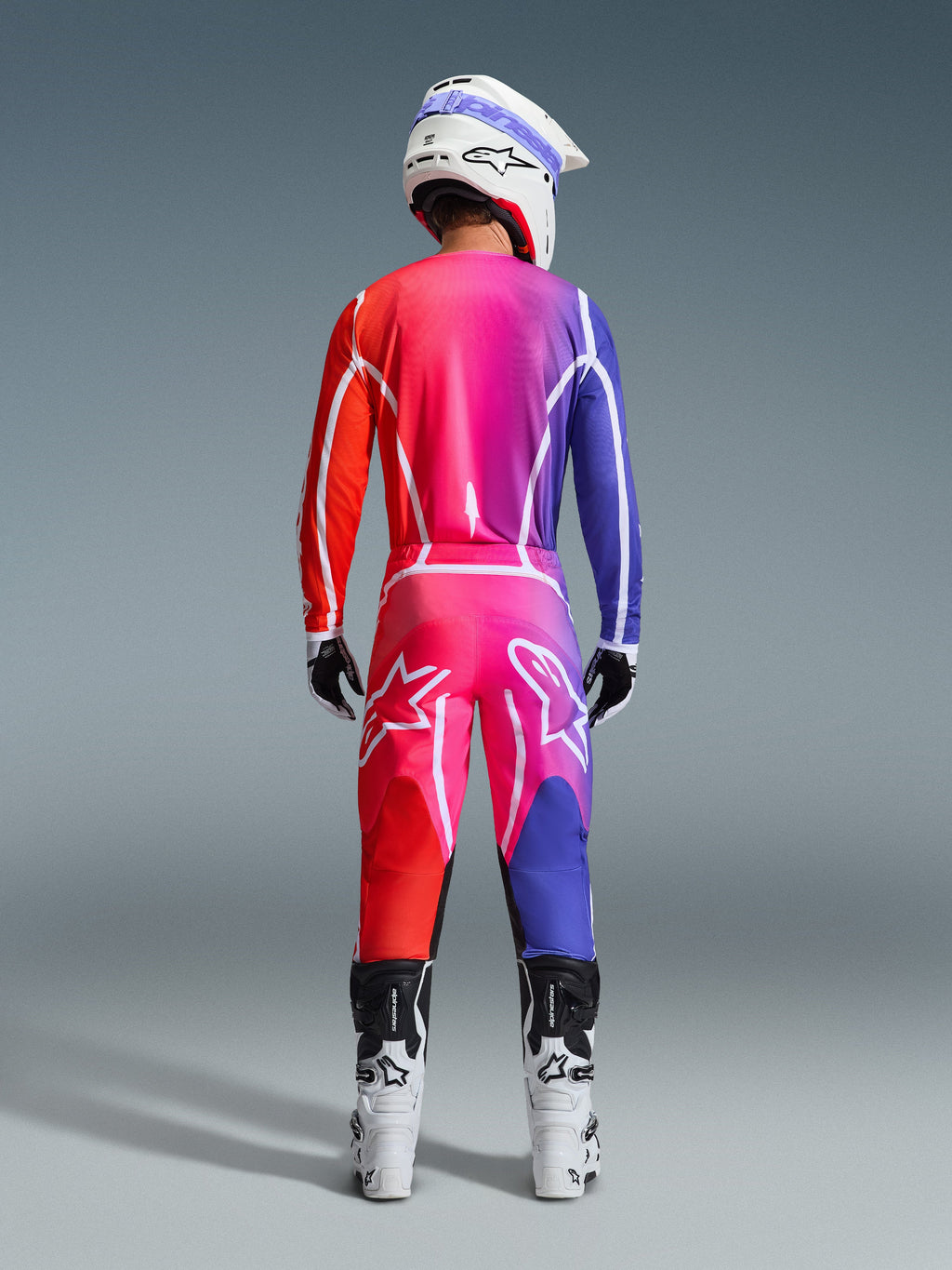 Set di equipaggiamento Alpinestars 2026 Fluid Apex, vista posteriore di un pilota di motocross che indossa maglia e pantaloni con una vivace sfumatura rosso, rosa e viola, linee grafiche bianche e grandi loghi a stella, abbinati a un casco bianco e stivali bianchi.