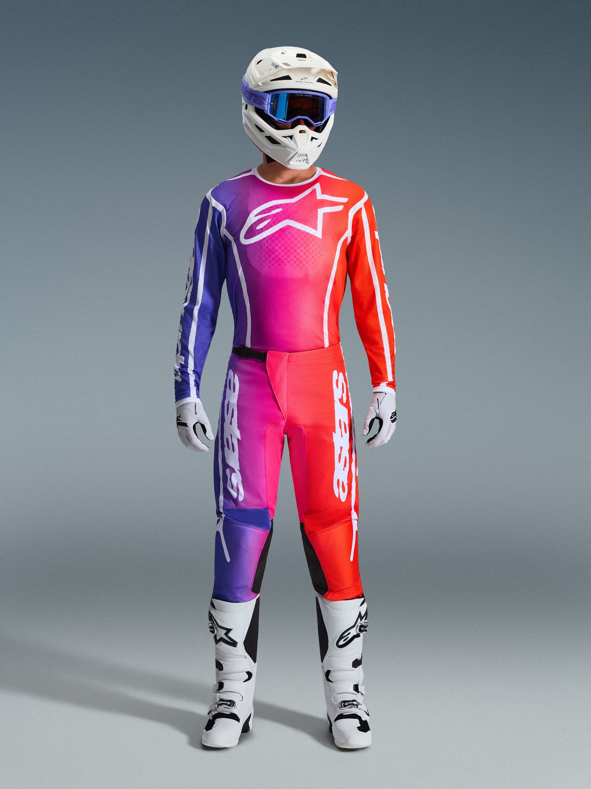 Pilota di motocross con un set completo di abbigliamento da gara Alpinestars 2026 Fluid Apex, caratterizzato da maglia e pantaloni con sfumature viola, rosa e arancione. L'outfit coordinato include un logo Alpinestars bianco sul petto, un casco bianco con occhiali blu e stivali bianchi, progettato per prestazioni fuoristrada.