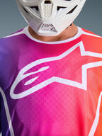 Maglia Alpinestars Fluid Apex 2026, maglia da motocross, sfumatura viola, rosa e arancione, grande logo a stella Alpinestars bianco sul petto, vista ravvicinata con mentoniera di un casco bianco