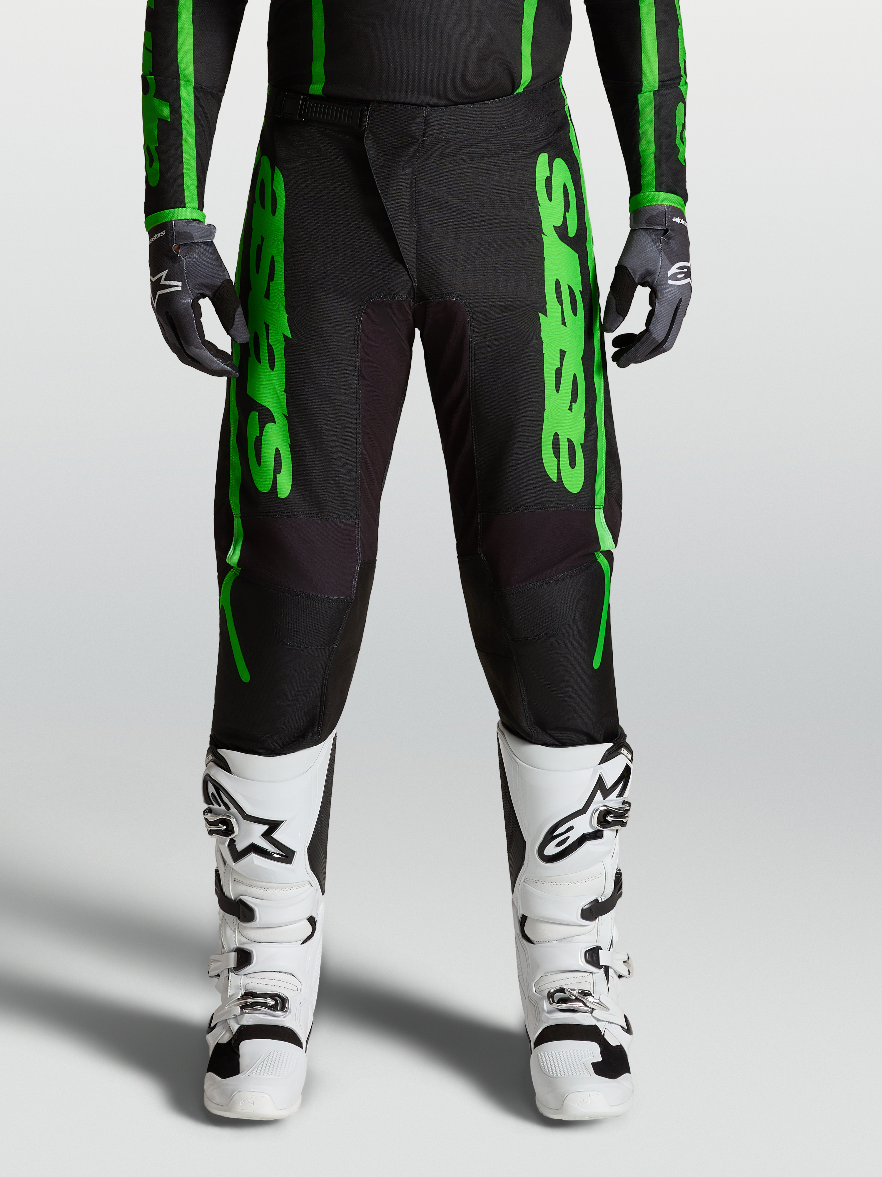 Alpinestars Pantaloni Fluid Apex V2, pantaloni motocross da uomo, neri e verdi, rinforzi su seduta e ginocchia, con stivali bianchi.