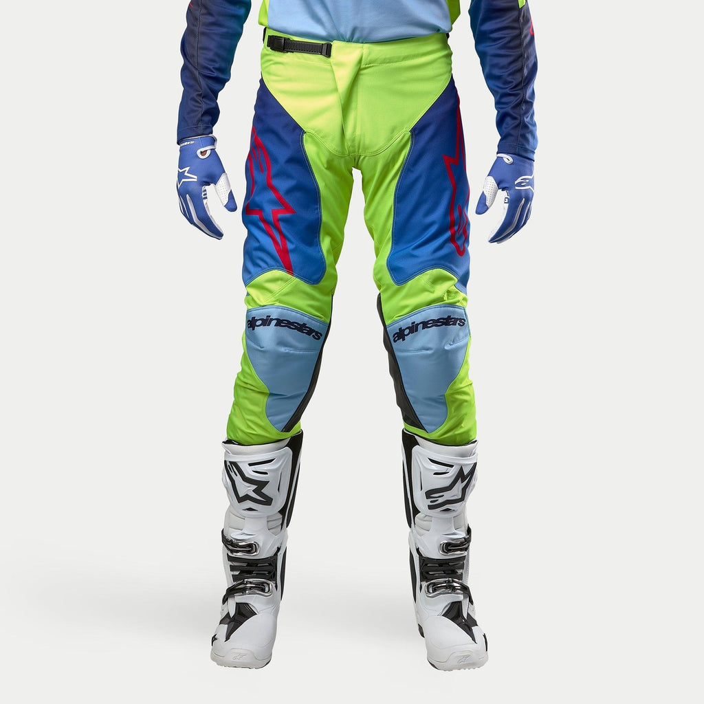 Alpinestars 2024 Racer Hoen Pants, pantaloni da motocross, Giallo Fluo e Blu Night Navy, indossati con stivali da motocross bianchi e neri e guanti blu, con loghi rossi vivaci, pannelli azzurri sulle ginocchia e un design ergonomico sportivo per la guida fuoristrada