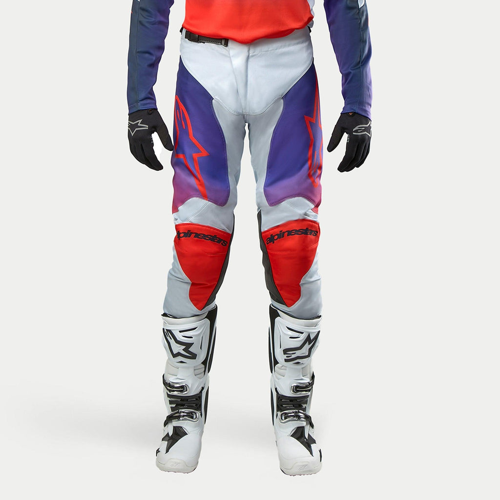Alpinestars 2024 Racer Hoen Pants, pantaloni da motocross in grigio chiaro con accenti arancione acceso e viola, logo stella rosso sulle cosce, pannelli protettivi sulle ginocchia, indossati con stivali bianchi e guanti neri.
