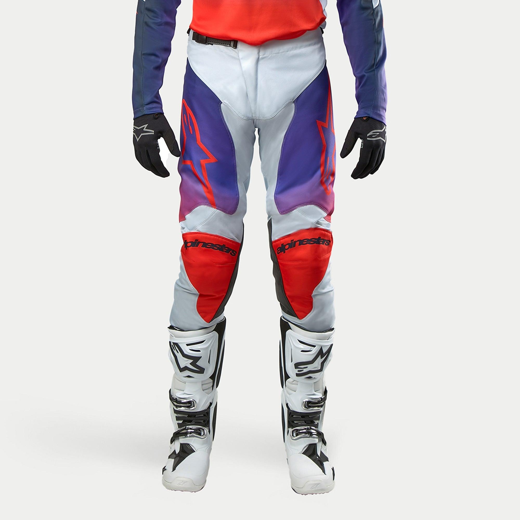 Alpinestars 2024 Racer Hoen Pants, pantaloni da motocross in grigio chiaro con accenti arancione acceso e viola, logo stella rosso sulle cosce, pannelli protettivi sulle ginocchia, indossati con stivali bianchi e guanti neri.