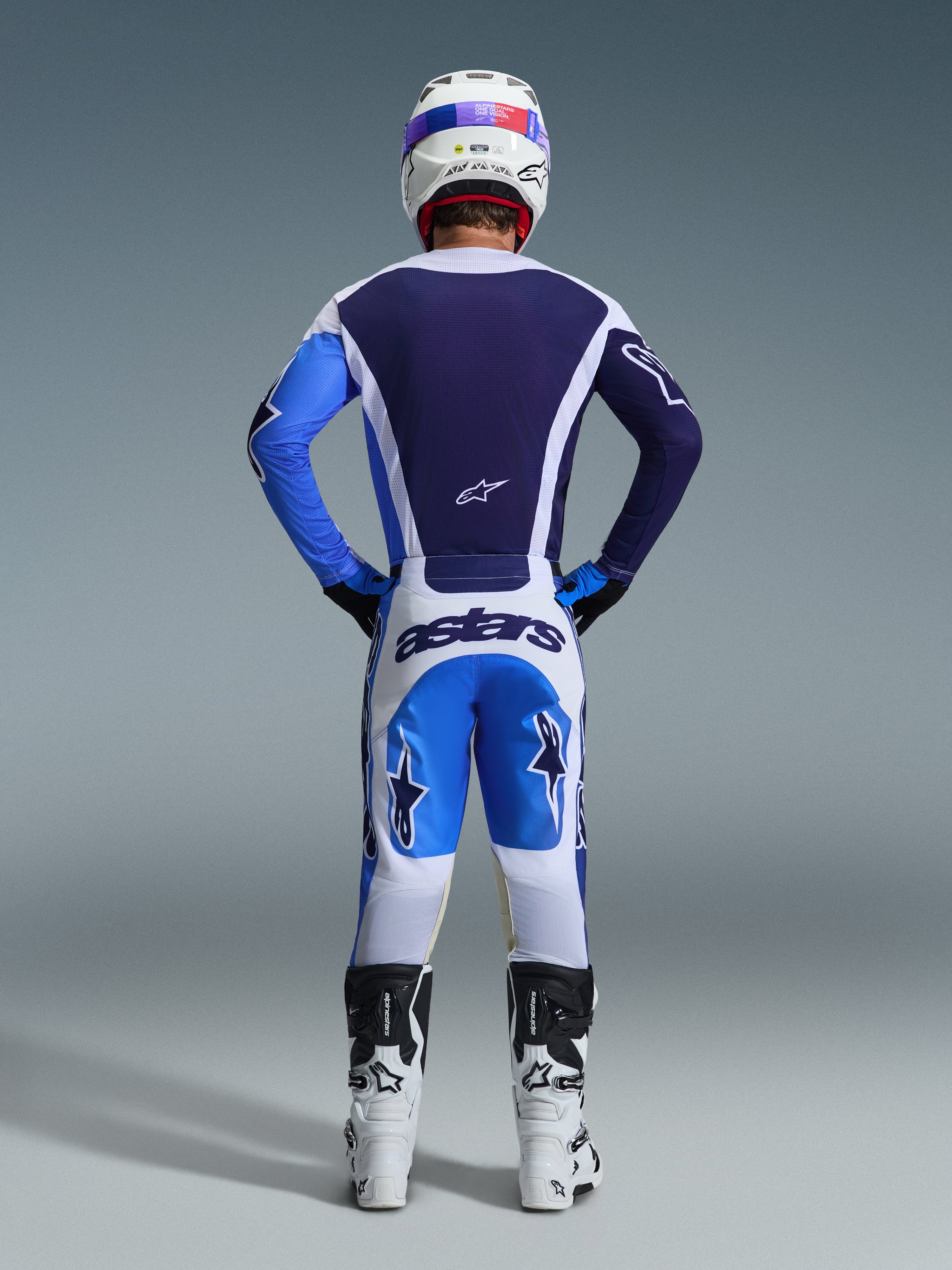Pilota di motocross visto da dietro con il set Alpinestars 2026 Racer Air Portl, maglia blu navy e azzurro con accenti bianchi, pantaloni bianchi con pannelli blu e marchio 'astars' navy, stivali da motocross bianchi e neri, casco da gara bianco, outfit coordinato per prestazioni fuoristrada