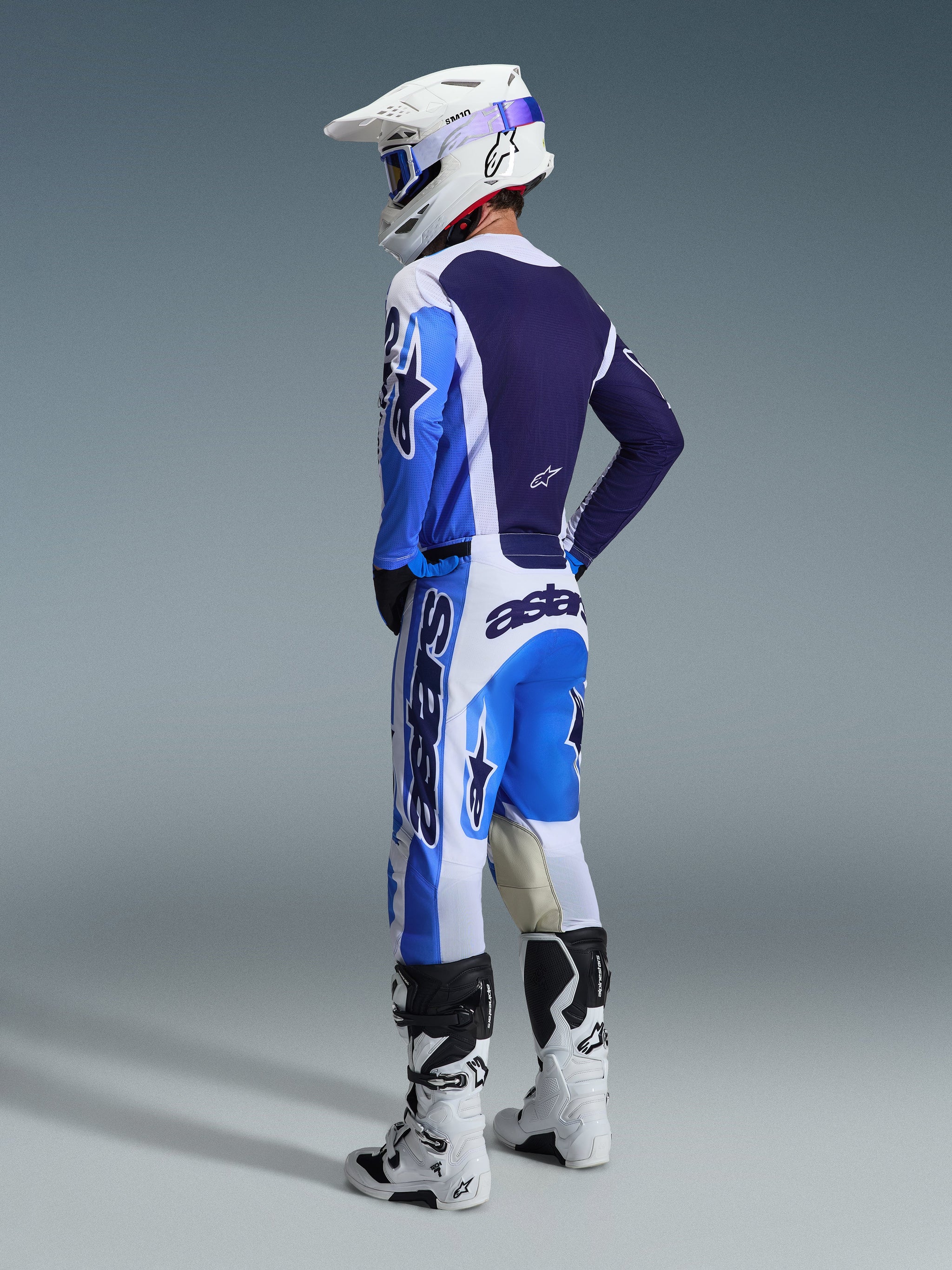 Pilota di motocross visto di spalle che indossa la maglia Alpinestars 2026 Racer Air Portl in blu Ucla e bianco, abbinata a pantaloni da motocross coordinati, casco Supertech bianco e stivali bianchi, che mostra un set completo di abbigliamento da gara coordinato.