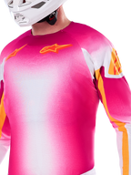 Alpinestars Limited Edition Supertech AFD MIG Jersey, maglia da motocross con sfumatura rosa fluo e bianco, loghi Astars e accenti sulle maniche arancioni, indossata da un pilota con casco bianco, vestibilità atletica da gara
