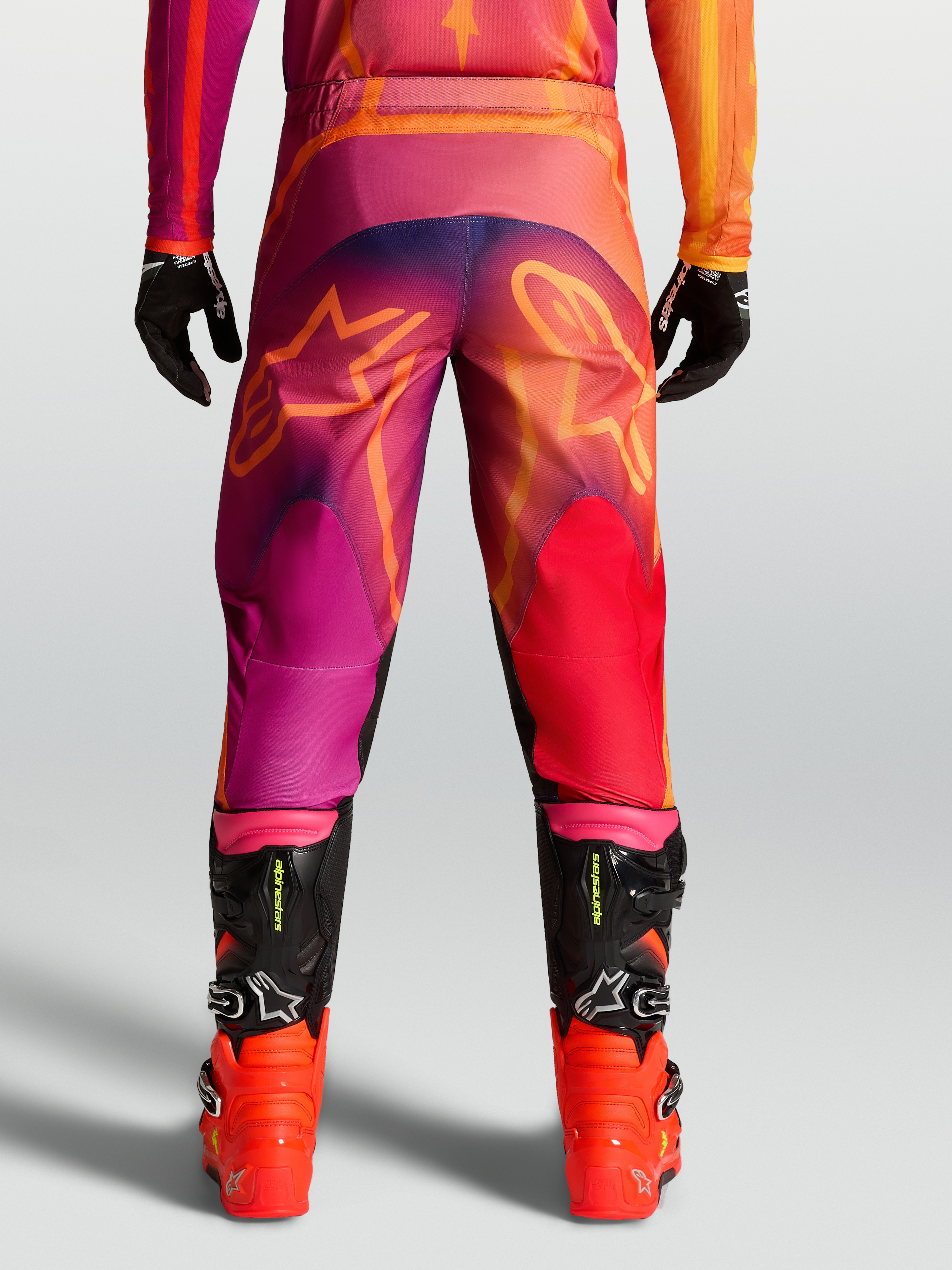 Alpinestars Pantaloni Fluid Kepler, pantaloni motocross da uomo, viola e arancione, con grafiche a stella posteriori e stivali neri.