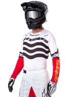 Pilota di motocross che indossa la maglia Alpinestars Limited Edition Techstar Deep, maglia bianca con strisce ondulate nere e parte inferiore delle maniche rossa, abbinata a casco nero, occhiali a specchio, guanti bianchi e neri e pantaloni da corsa bianchi, completo da motocross ad alte prestazioni.