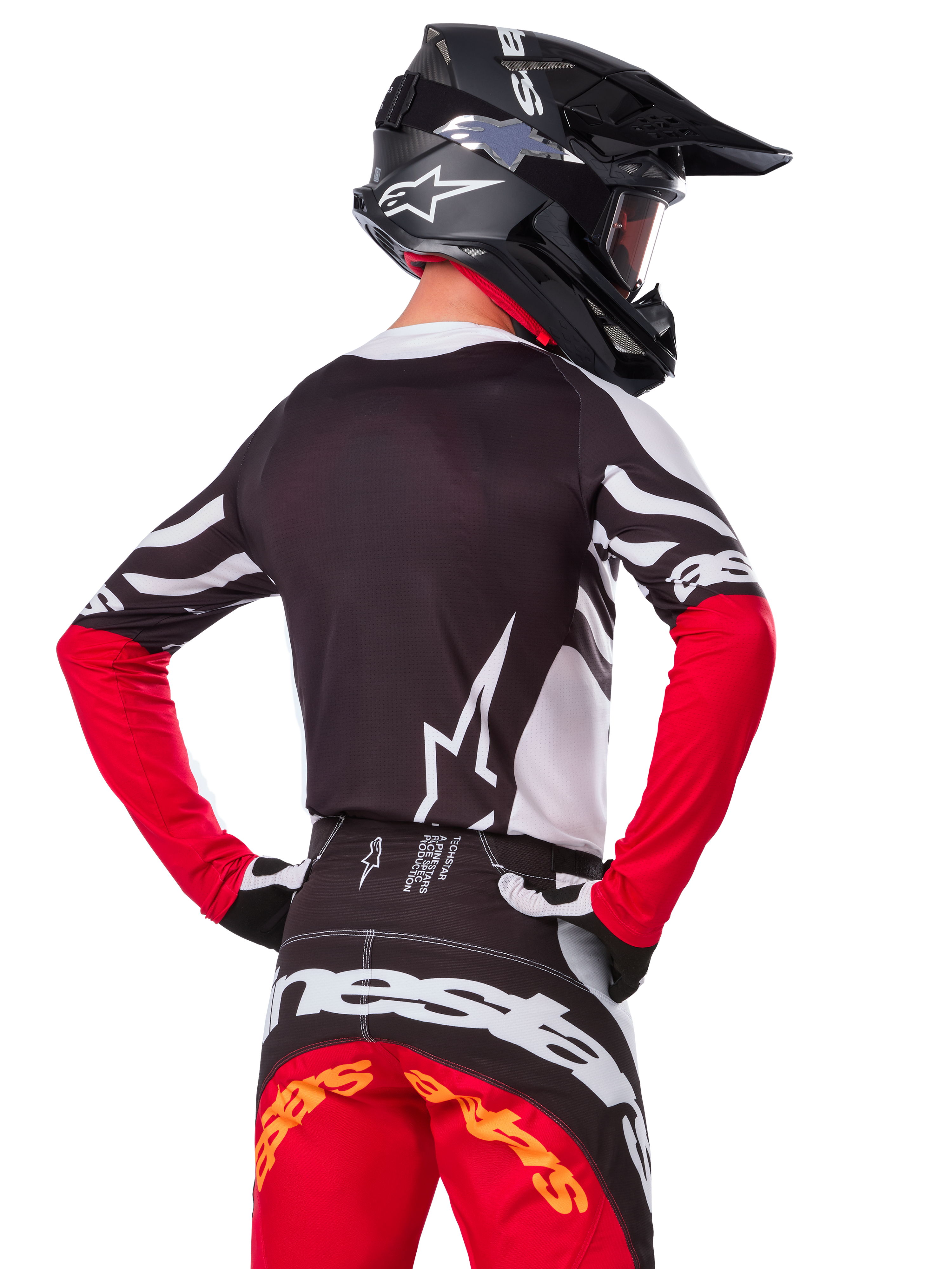 Pilota di motocross con abbigliamento completo da gara, maglia Alpinestars Limited Edition Techstar Deep in nero, bianco e rosso acceso, vista posteriore, pantaloni Techstar Deep coordinati, casco nero con maschera, guanti bianchi e neri, completo professionale per fuoristrada