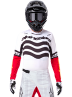 Pilota di motocross con abbigliamento completo da gara, maglia Limited Edition Techstar Deep bianca e nera con maniche rosse, grafiche ondulate e marchio Alpinestars, pantaloni bianchi, casco e maschera neri, outfit coordinato per corse off-road