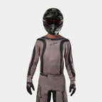 Pilota di motocross che indossa la maglia Alpinestars Fluid Lurv 2024 in Mud Black, un tessuto grigio-marrone terroso con strisce grafiche nere e logo a stella sul petto, abbinata a un casco nero, occhiali rossi e guanti neri per un look da corsa off-road coordinato