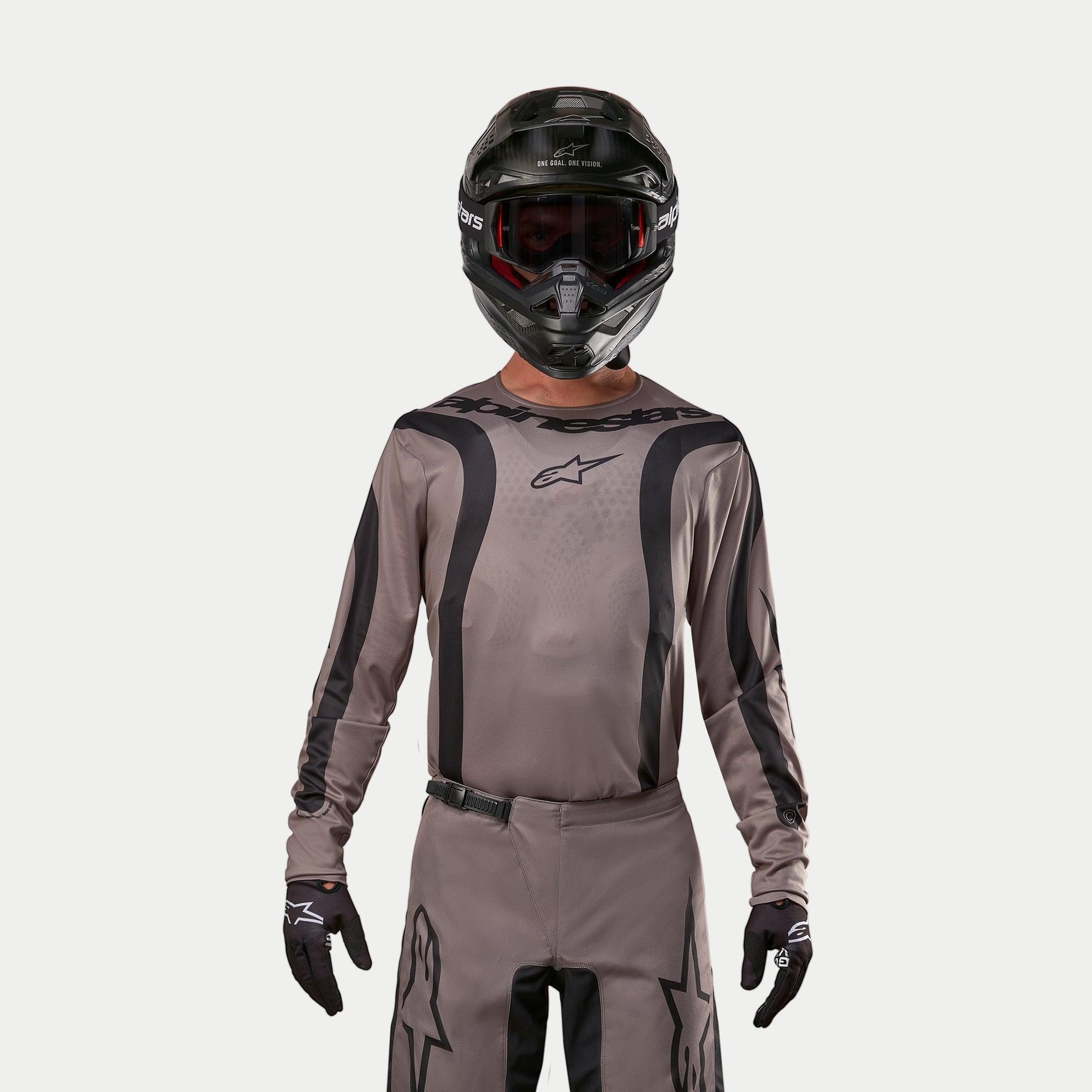 Pilota di motocross che indossa la maglia Alpinestars Fluid Lurv 2024 in Mud Black, un tessuto grigio-marrone terroso con strisce grafiche nere e logo a stella sul petto, abbinata a un casco nero, occhiali rossi e guanti neri per un look da corsa off-road coordinato
