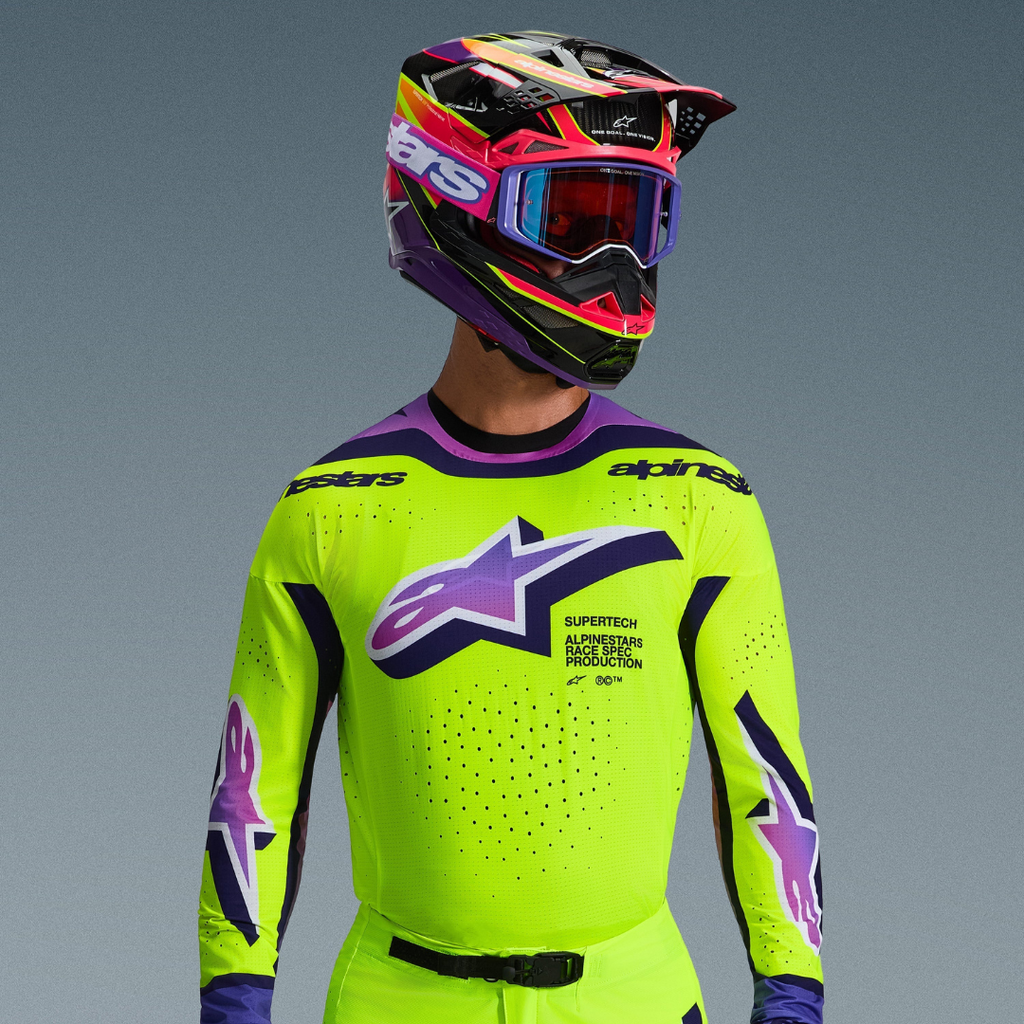 Pilota di motocross con completo Alpinestars Supertech Vista 2026, maglia giallo neon con dettagli viola e neri, grande logo viola sfumato sul petto, casco multicolore vivace coordinato e occhiali viola, con ventilazione tagliata al laser e vestibilità atletica professionale