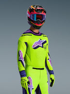 Pilota di motocross con maglia Alpinestars Supertech Vista 2026 e completo coordinato, giallo acceso con accenti viola e neri, grande logo Astar viola sul petto e sulle maniche, guanti coordinati e casco da corsa multicolore con maschera, vestibilità atletica professionale per competizioni fuoristrada.