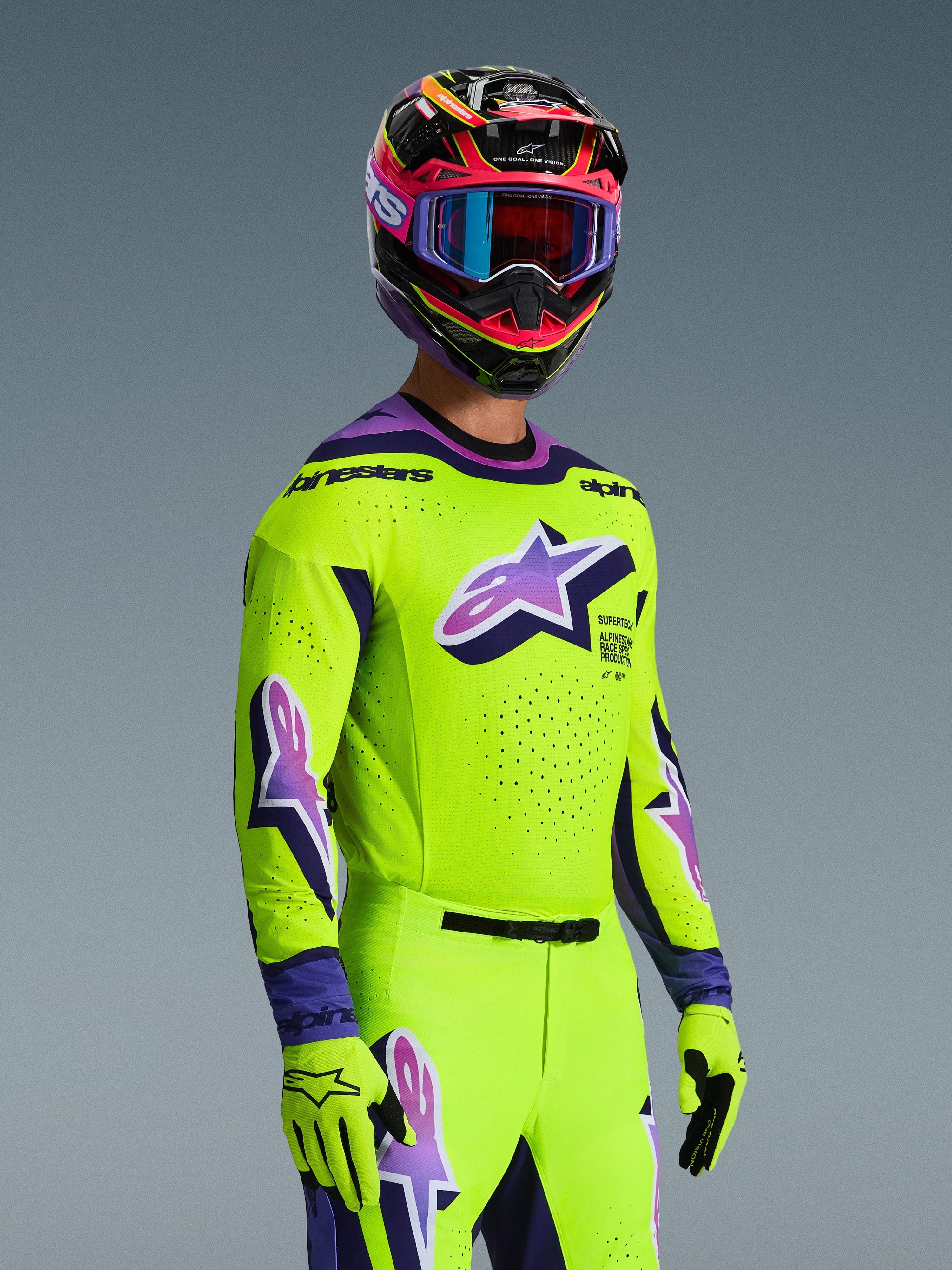 Pilota di motocross con maglia Alpinestars Supertech Vista 2026 e completo coordinato, giallo acceso con accenti viola e neri, grande logo Astar viola sul petto e sulle maniche, guanti coordinati e casco da corsa multicolore con maschera, vestibilità atletica professionale per competizioni fuoristrada.