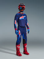 Pilota di motocross con equipaggiamento completo da gara, maglia Alpinestars 2026 Supertech Vista in Dark Navy, Hot Coral e Cobalt, pantaloni coordinati con grandi loghi a stella, stivali da motocross rosso acceso, guanti blu e casco multicolore con occhiali rossi, set professionale per fuoristrada