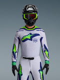 Alpinestars 2026 Supertech Vista Jersey, maglia da motocross, grigio chiaro, giallo fluo e blu, indossata da un pilota con pantaloni coordinati e casco nero, con un grande logo sul petto e perforazioni laser per la ventilazione, vestibilità atletica di livello professionale