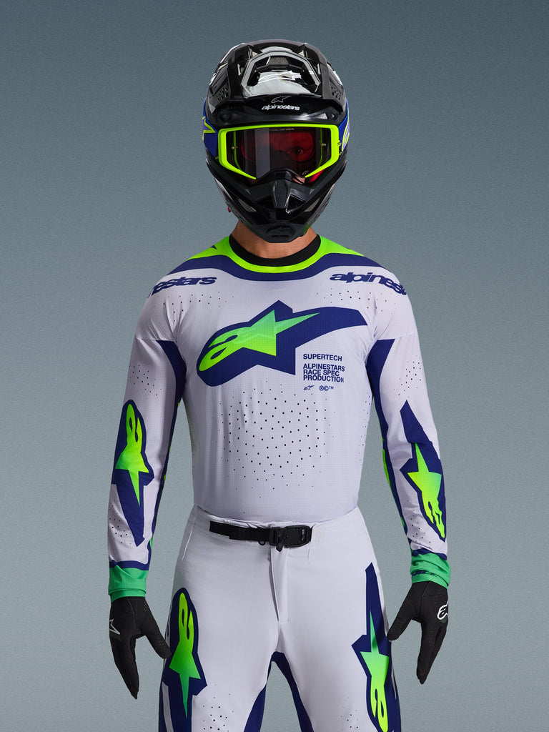 Alpinestars 2026 Supertech Vista Jersey, maglia da motocross, grigio chiaro, giallo fluo e blu, indossata da un pilota con pantaloni coordinati e casco nero, con un grande logo sul petto e perforazioni laser per la ventilazione, vestibilità atletica di livello professionale