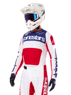 Pilota di motocross con maglia Alpinestars Limited Edition Techstar Fortitude, rosso acceso, bianco e blu, con casco bianco, occhiali blu e pantaloni coordinati, abbigliamento da gara ad alte prestazioni per competizioni off-road