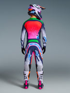 Pilota di motocross visto dal retro con il completo Alpinestars Limited Edition Supertech Records, maglia e pantaloni multicolore con design splatter rosa, arancione e blu, casco coordinato, guanti neri e stivali bianchi e rosa, abbigliamento professionale per fuoristrada.