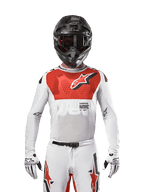 Pilota di motocross con maglia Alpinestars 2024 Supertech Ward in White Hot Orange, maglia in rete elastica bianca con grafica e logo rossi sul petto, casco nero con occhiali scuri, guanti bianchi, abbigliamento professionale per performance off-road