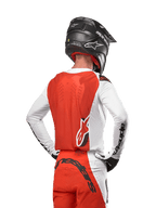 Pilota di motocross con maglia Alpinestars Supertech Ward 2024 in White Hot Orange, vista posteriore con pannello posteriore arancione e maniche bianche con branding Alpinestars nero, abbinata a pantaloni coordinati e casco nero, attrezzatura professionale per corse fuoristrada