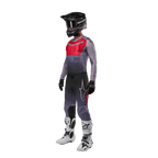 Pilota di motocross con abbigliamento completo da gara, maglia Alpinestars 2024 Supertech Dade in Iron Red Berry e grigio, pantaloni grigi coordinati con accenti rossi, casco nero con occhiali, stivali bianchi e neri, guanti neri, outfit coordinato per corse fuoristrada
