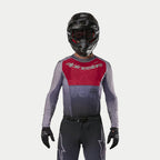 Pilota di motocross che indossa la maglia Alpinestars Supertech Dade 2024 in Iron Red Berry, caratterizzata da un pannello rosso sul petto e maniche grigie con branding argento, abbinata a pantaloni coordinati, casco nero e guanti neri, equipaggiamento professionale da gara fuoristrada