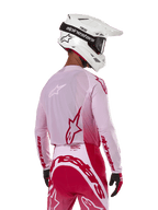 Pilota di motocross visto di spalle che indossa la maglia Alpinestars Supertech Dade 2024 in Red Berry Lilac, con pannelli in rete traspirante e loghi Alpinestars rossi, abbinata a pantaloni coordinati rossi e lilla e un casco Alpinestars bianco con maschera, abbigliamento professionale da gara off-road