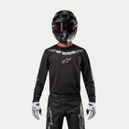 Maglia Alpinestars Fluid Graphite 2024, maglia da motocross nero e argento, indossata da un pilota con casco nero e occhiali rossi, con logo Alpinestars argento sul petto, coordinata con pantaloni neri e guanti bianchi per prestazioni fuoristrada