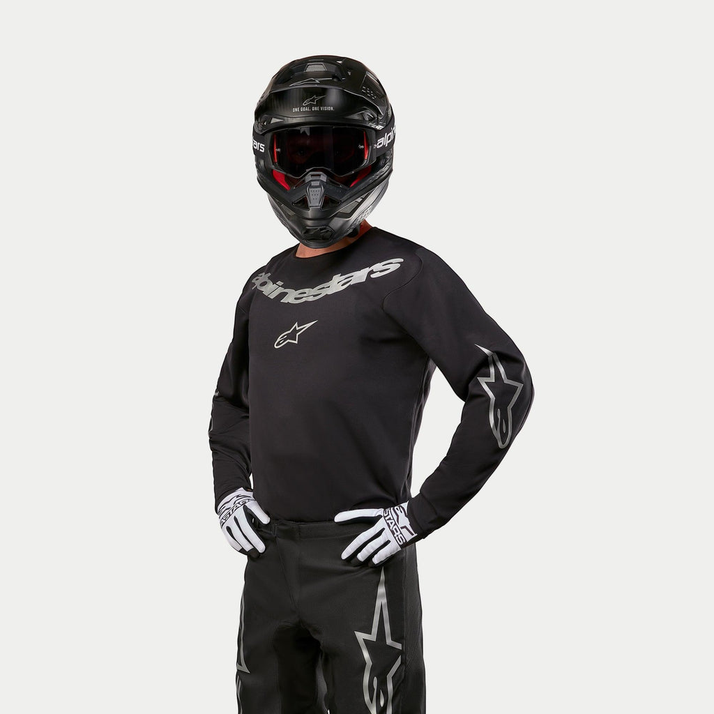 Pilota di motocross che indossa la maglia Alpinestars Fluid Graphite 2024, nero e argento, abbinata a pantaloni da motocross neri, casco nero con occhiali con lenti rosse e guanti bianchi e neri, con loghi del marchio argentati sul petto e sulle maniche.