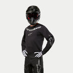 Pilota di motocross che indossa la maglia Alpinestars Fluid Graphite 2024, nero e argento, abbinata a pantaloni da motocross neri, casco nero con occhiali con lenti rosse e guanti bianchi e neri, con loghi del marchio argentati sul petto e sulle maniche.
