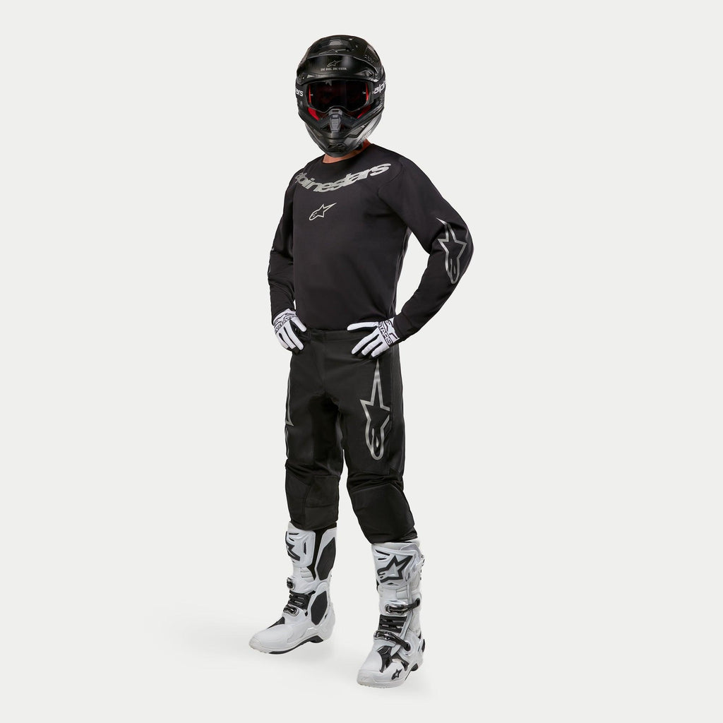 Pilota di motocross con maglia Alpinestars 2024 Fluid Graphite in nero e argento, pantaloni neri coordinati con loghi a stella argento, stivali e guanti bianchi e neri, e casco nero con occhiali rossi, abbigliamento completo da gara off-road